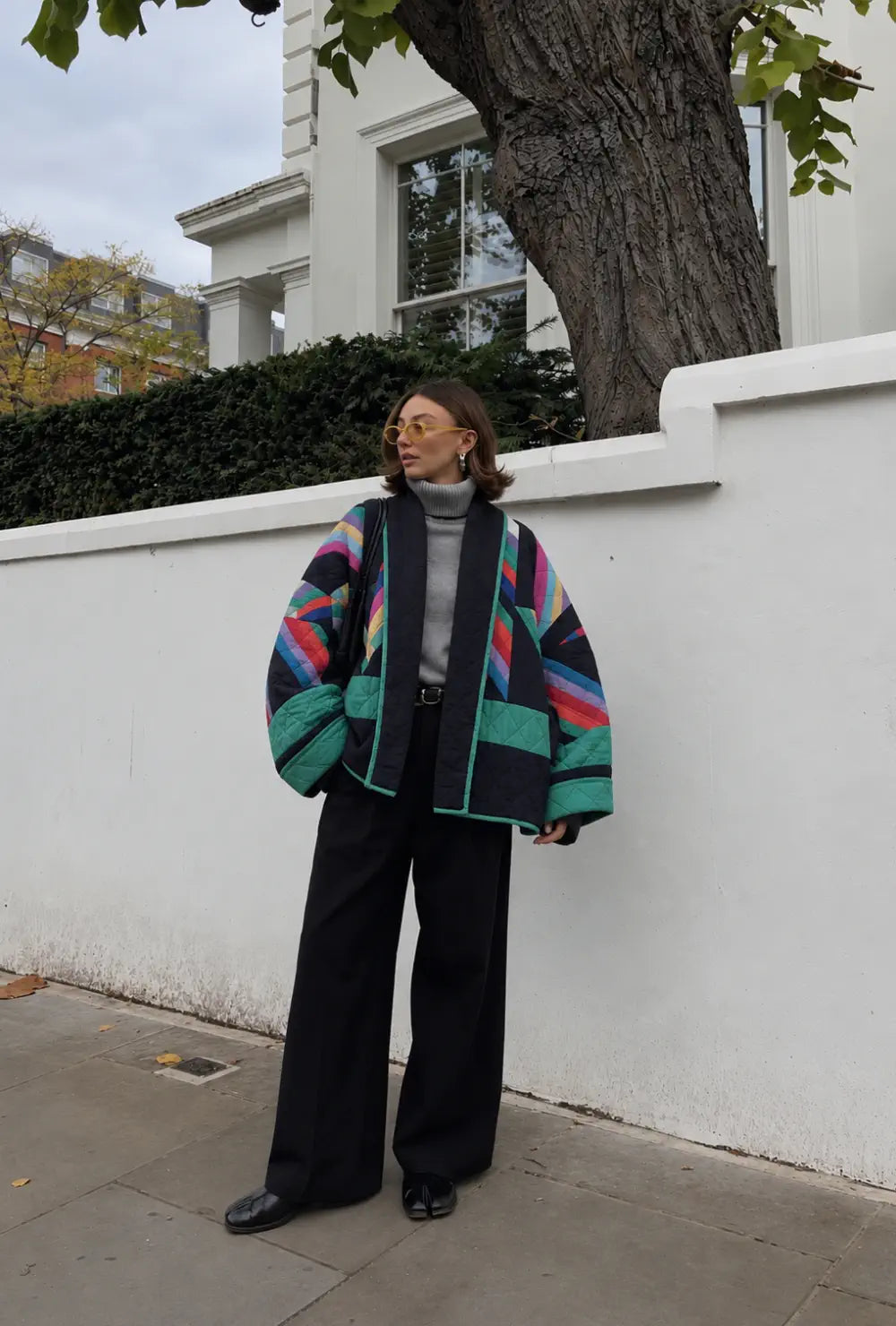 Blazer kimono femme matelassé style années 80 porté sur un col roulé gris et un pantalon noir devant un mur blanc