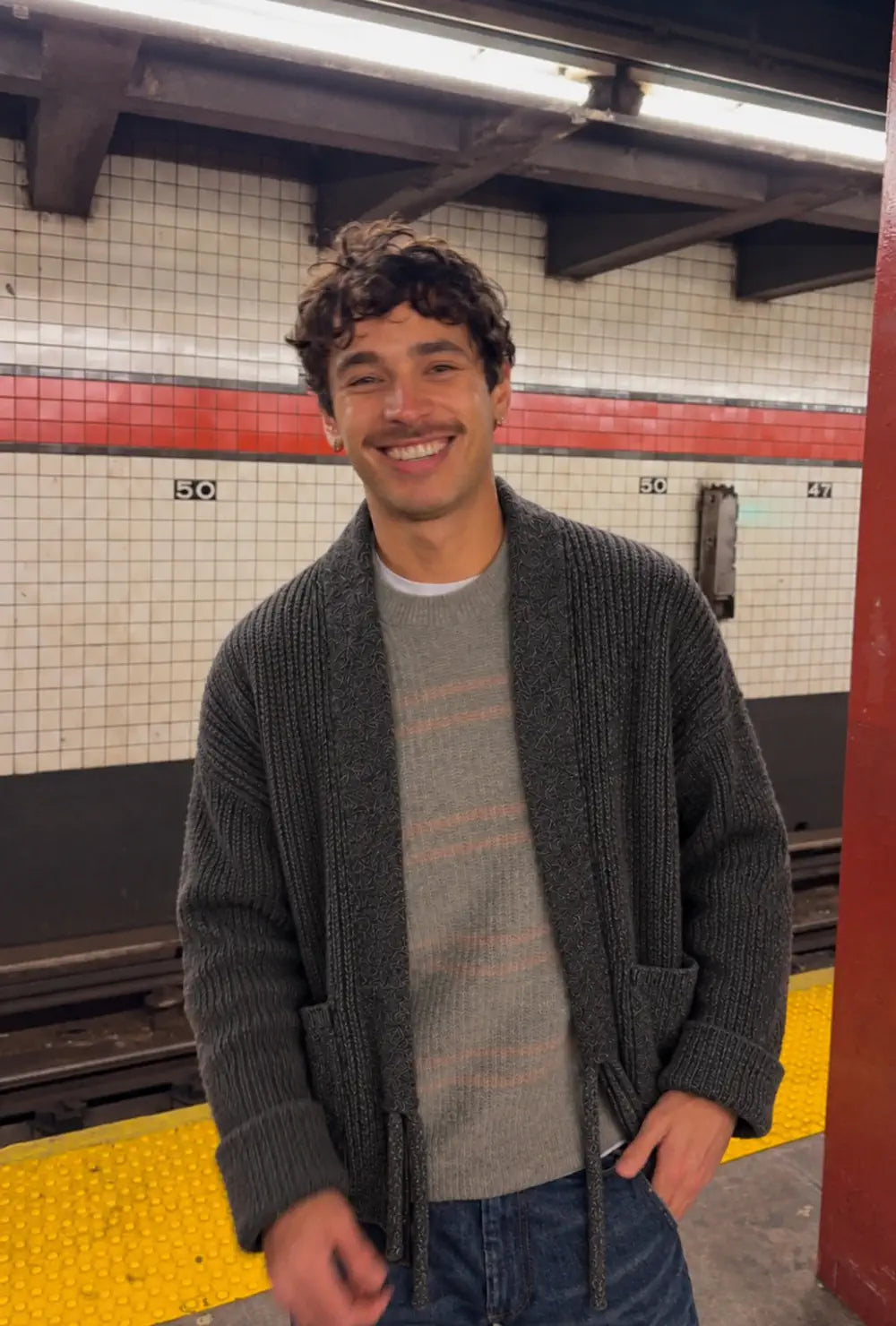 Cardigan kimono homme gris en laine oversize porté ouvert sur un pull rayé dans une station de métro