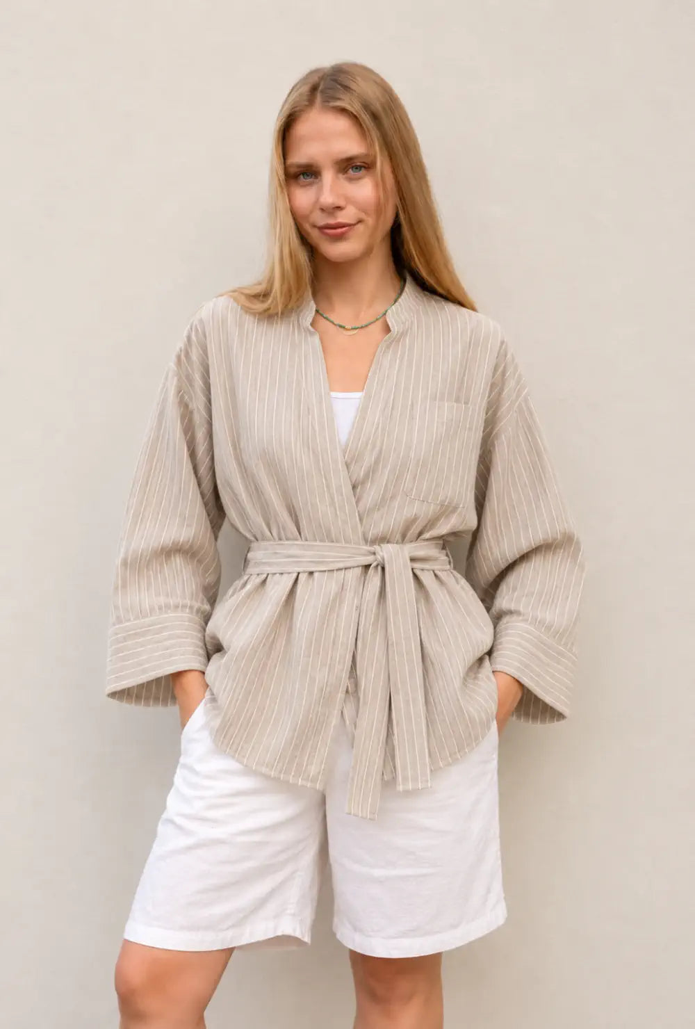 Kimono chemise femme beige en coton rayé oversize porté ceinturé sur un short blanc devant un mur clair
