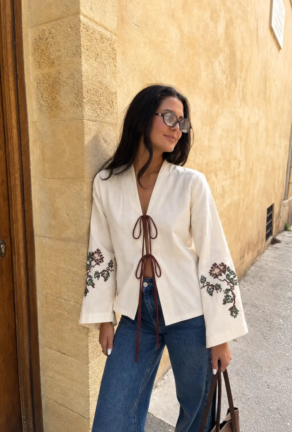 Kimono femme beige brodé à motif fleuri porté noué sur un jean bleu contre un mur ocre en ville