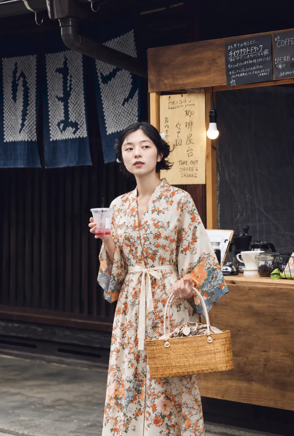 Kimono long femme beige à motifs fleuris bohème porté ceinturé avec un panier en osier devant un stand de café