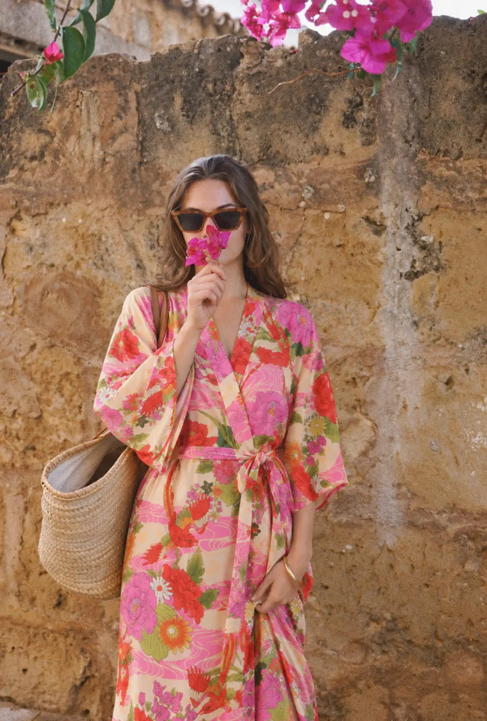 Kimono long femme rose pastel fleuri porté ceinturé avec un panier en osier devant un vieux mur en pierre