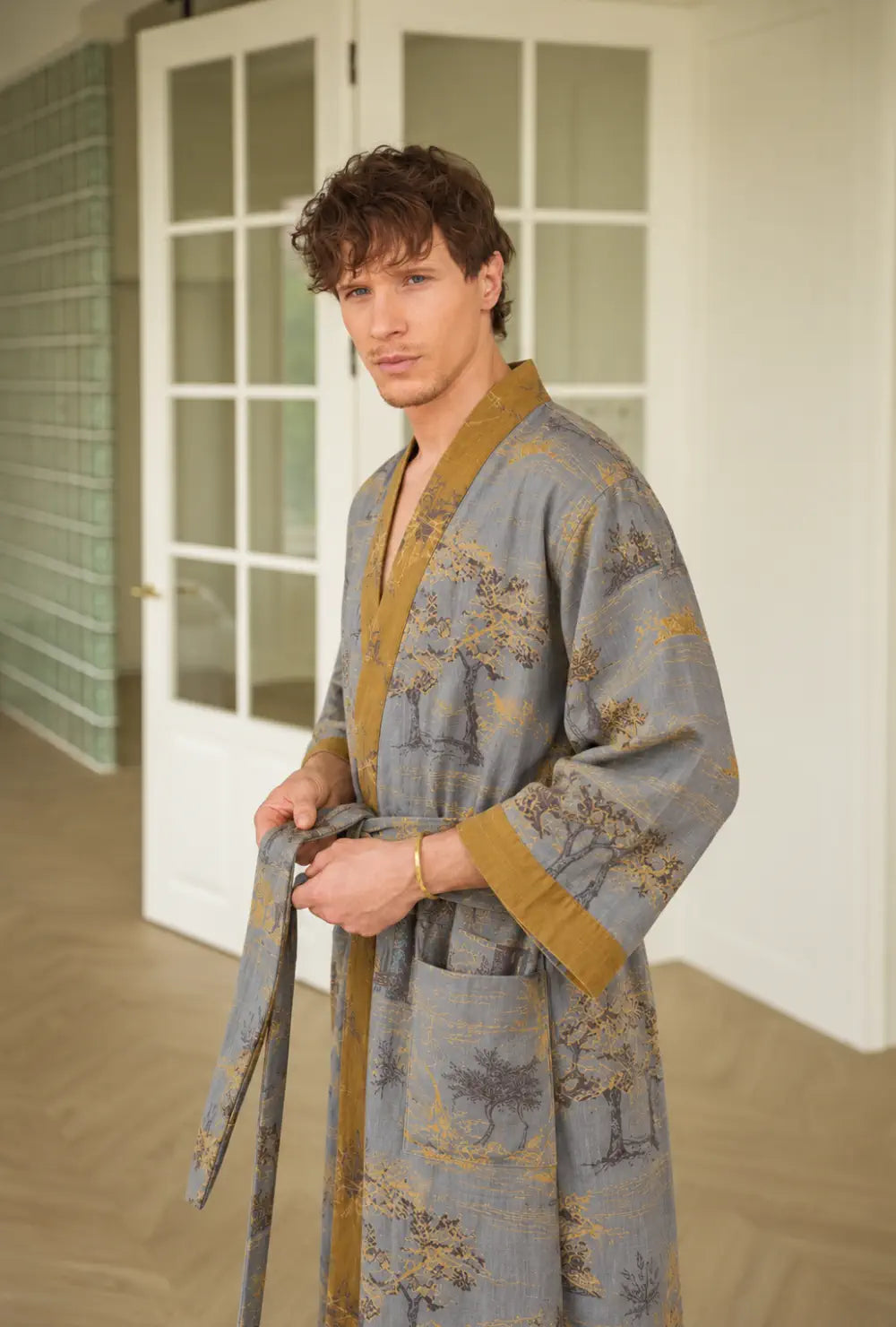 Peignoir japonais homme gris en coton à col V porté en nouant la ceinture devant une porte vitrée blanche