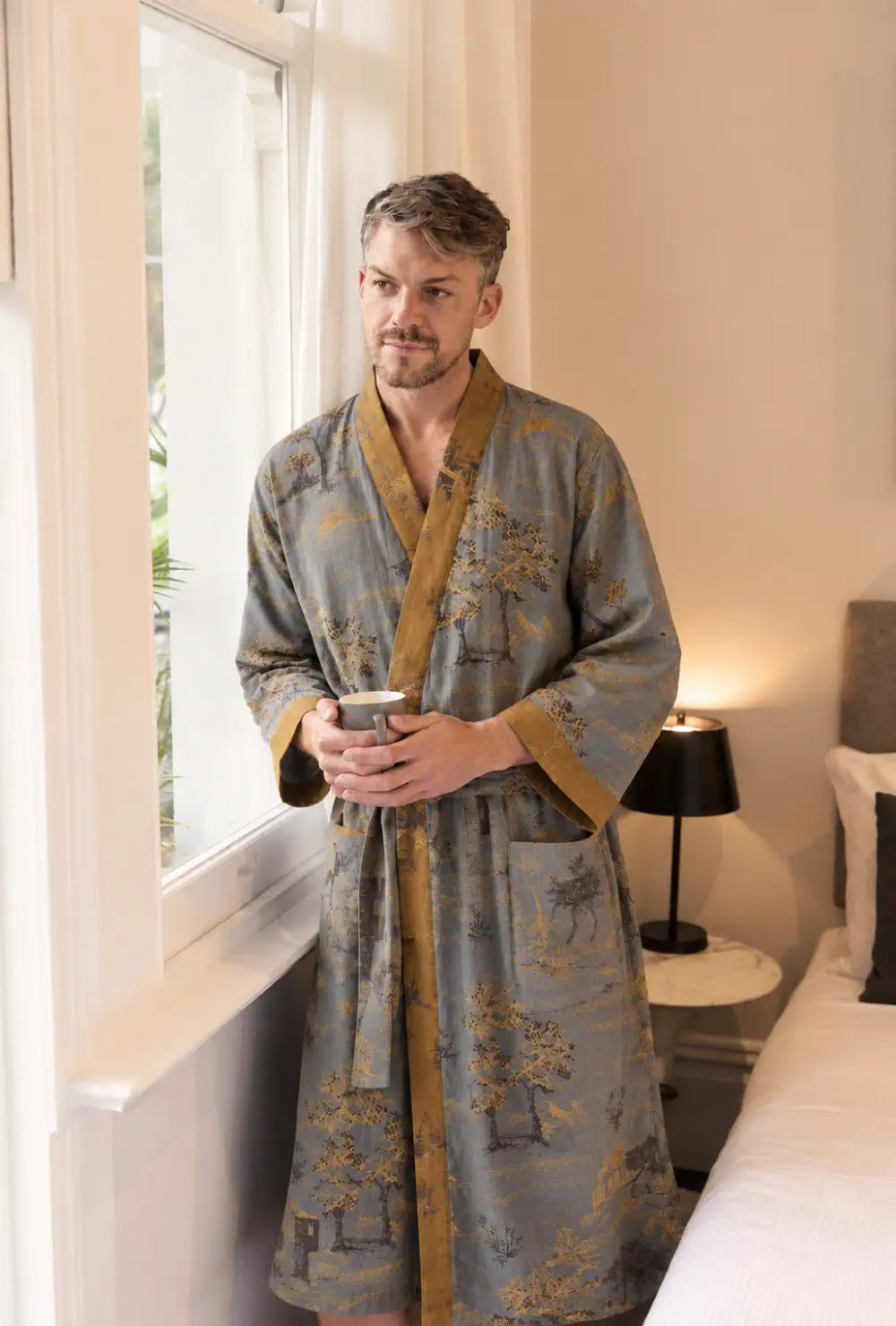 Peignoir japonais homme gris en coton à col V porté ceinturé avec un mug près de la fenêtre d'une chambre