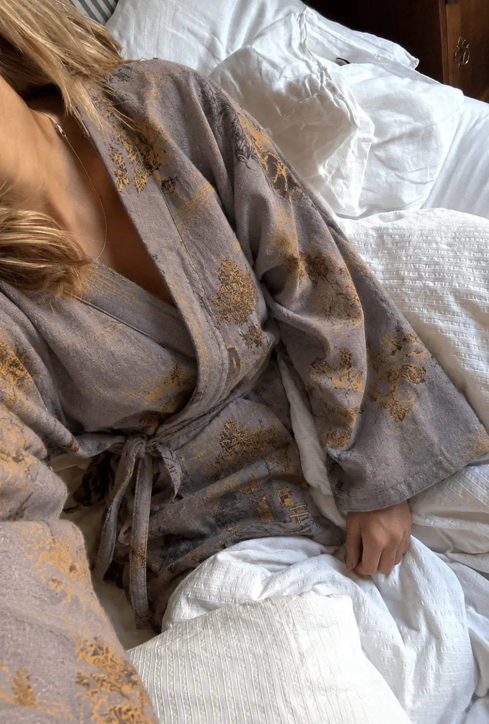 Peignoir kimono femme gris en coton porté noué à la taille dans un lit aux draps blancs