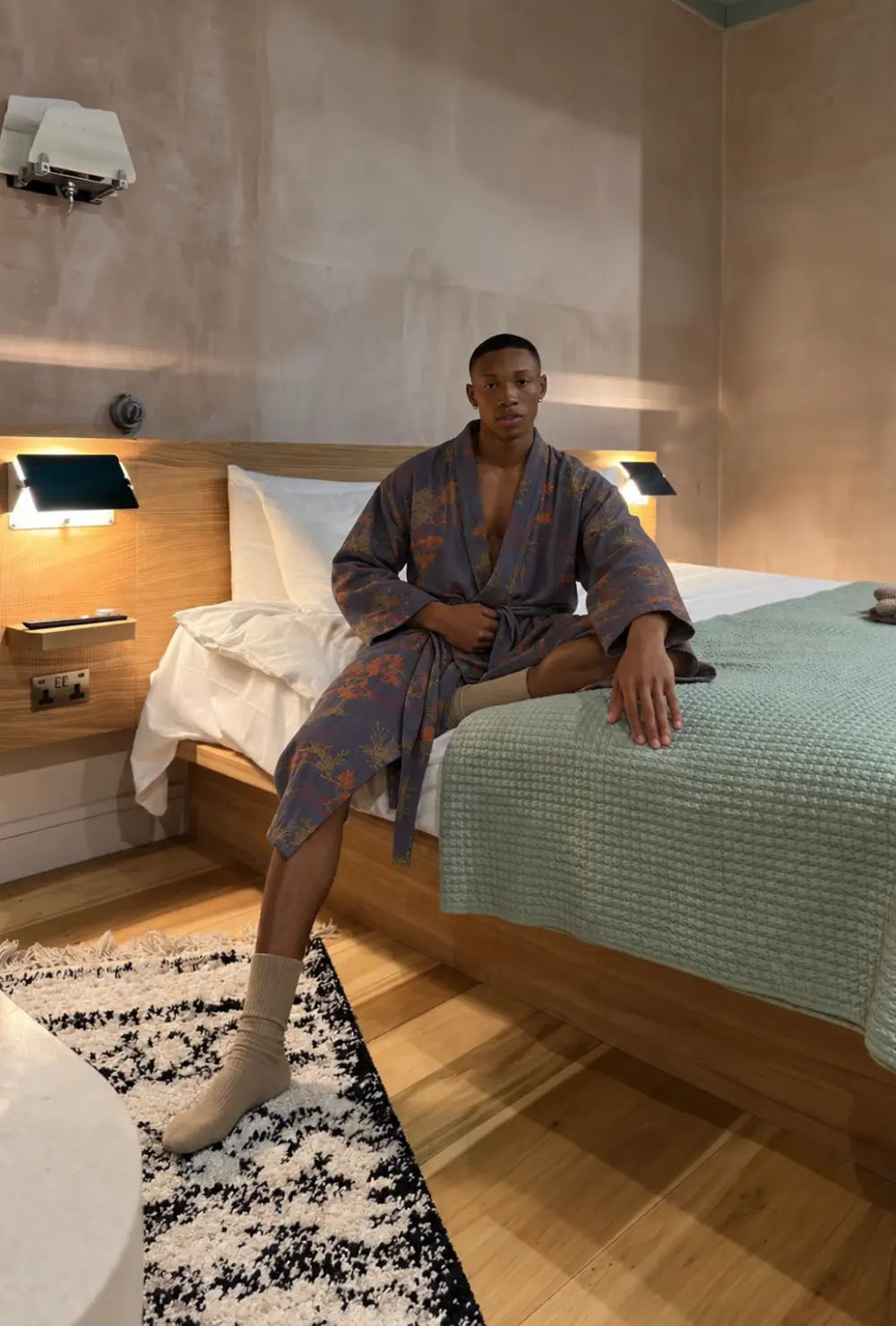 Peignoir kimono homme bleu en coton à motif japonais porté ceinturé, assis au bord d'un lit dans une chambre
