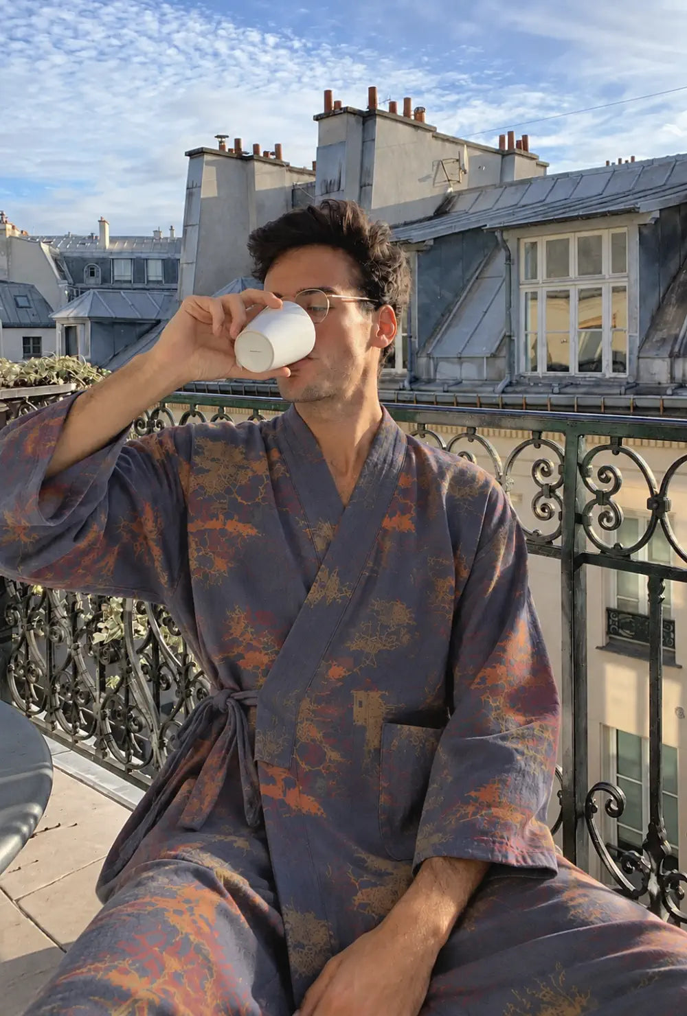 Peignoir kimono homme bleu en coton à motif japonais porté en buvant un café sur un balcon face aux toits