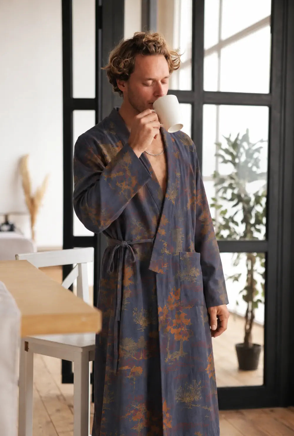 Peignoir kimono homme bleu en coton à motif japonais porté ceinturé, une tasse aux lèvres près d'une verrière