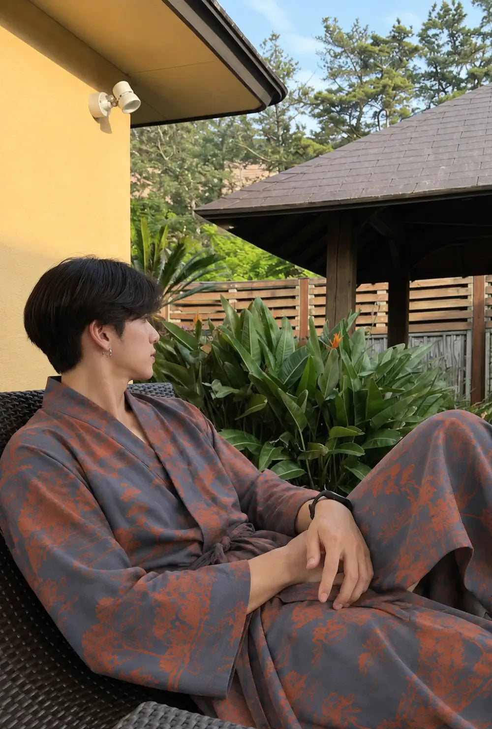 Peignoir kimono homme bleu en coton porté ceinturé, installé dans un fauteuil sur une terrasse arborée