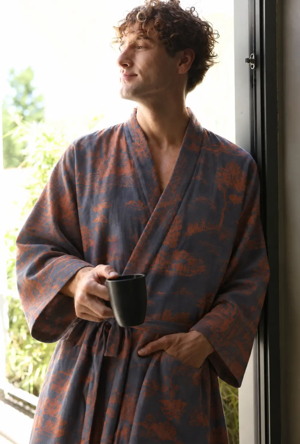 Peignoir kimono homme bleu en coton porté ceinturé avec un mug noir devant une fenêtre ouverte