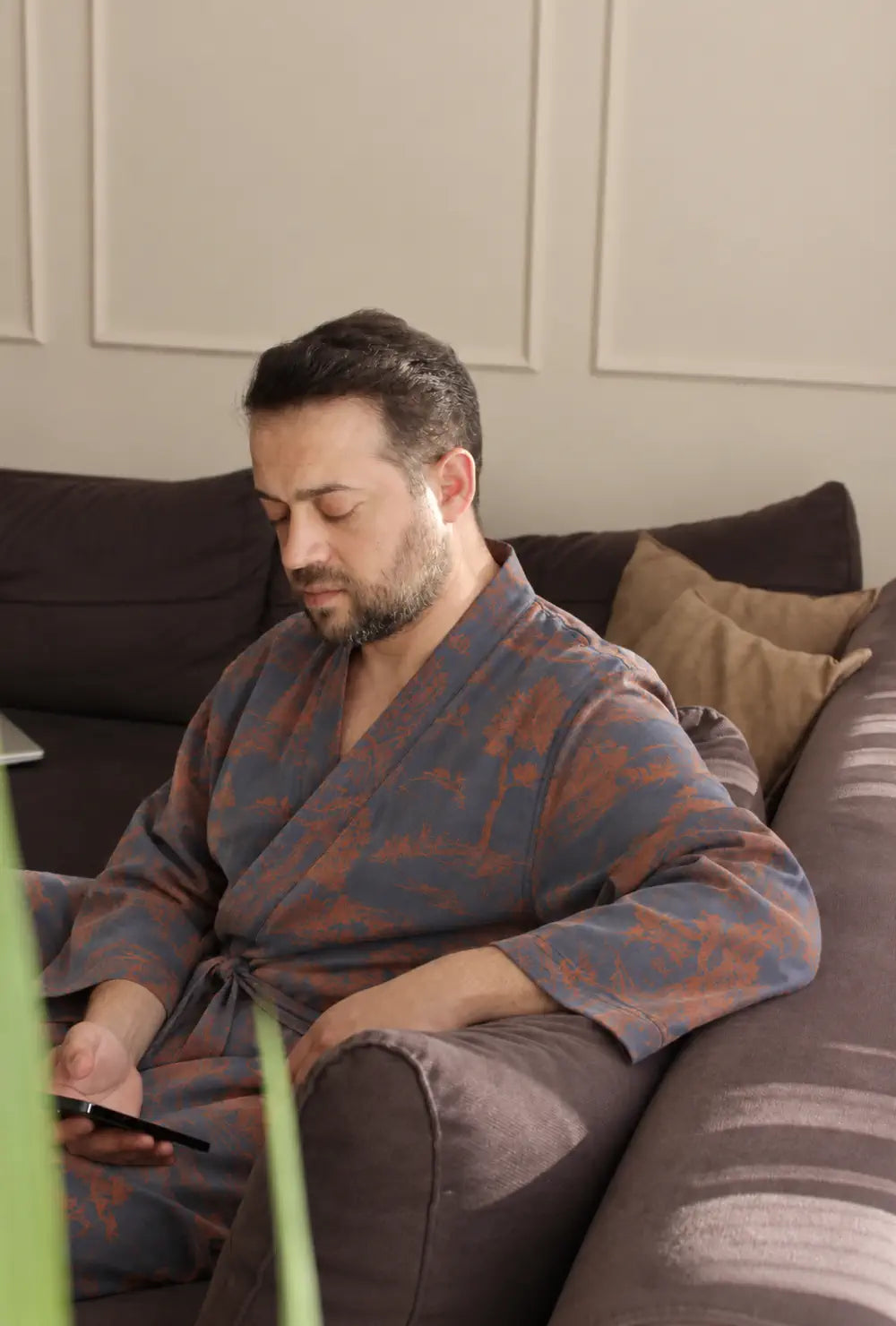Peignoir kimono homme bleu en coton porté ceinturé, assis sur un canapé avec un téléphone en main