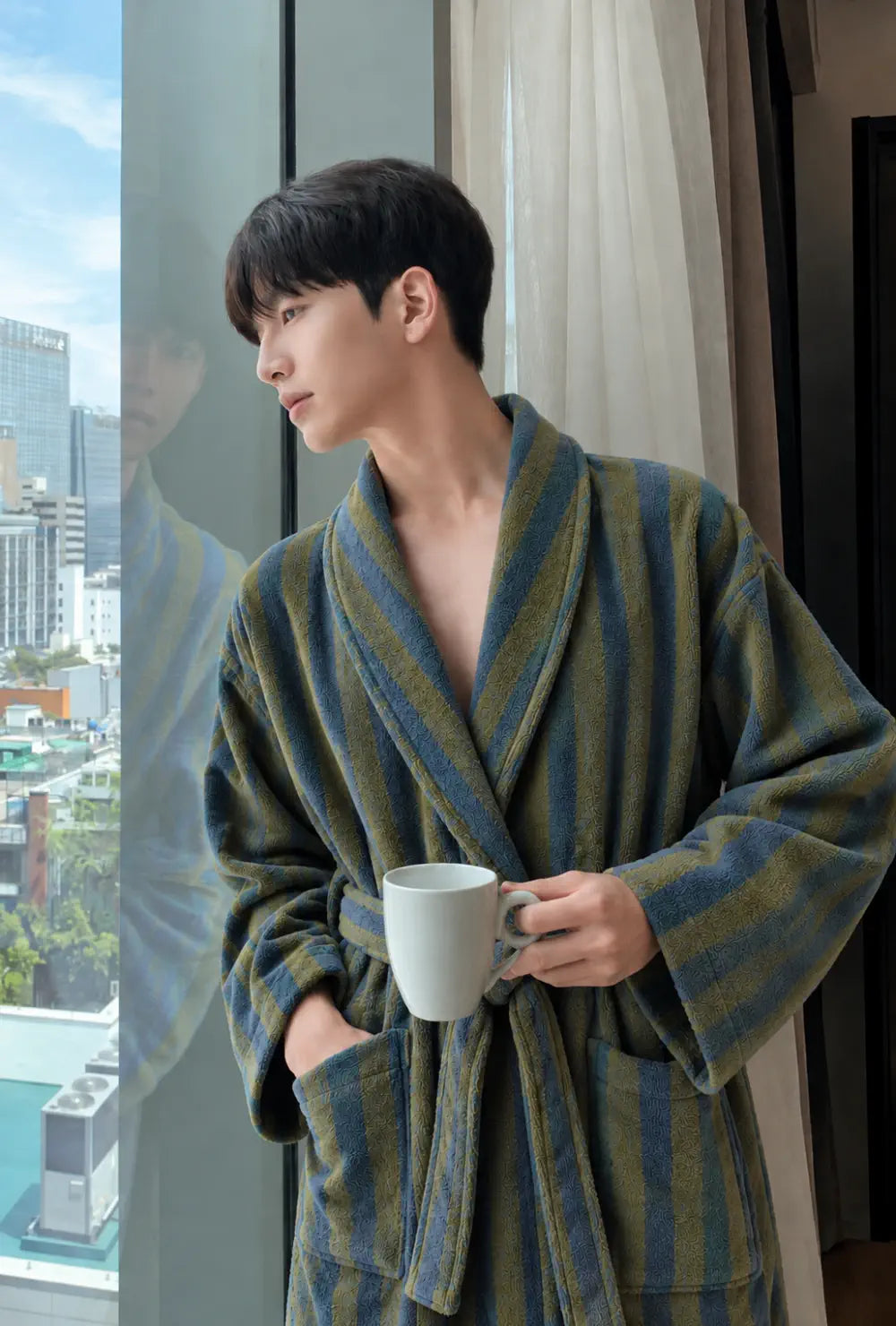 Peignoir kimono homme vert en polaire chaude porté ceinturé, une tasse à la main près d'une baie vitrée