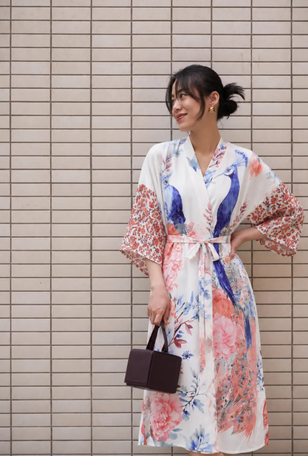 Robe japonaise femme blanche à motif paon et fleurs portée ceinturée avec un petit sac devant un mur carrelé