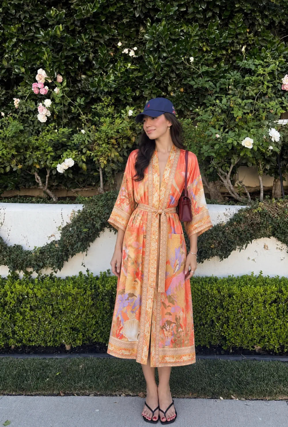 Robe japonaise femme orange fleurie portée ceinturée avec une casquette devant une haie de rosiers