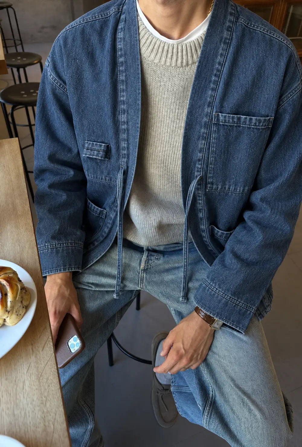 Veste kimono denim homme bleue en coton portée ouverte sur un pull gris et un jean dans un café