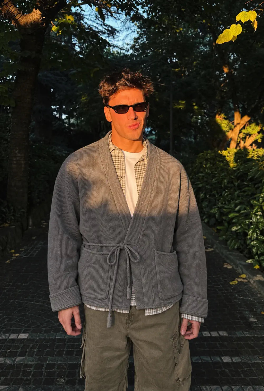 Veste kimono homme grise style cardigan portée nouée sur une chemise à carreaux dans une allée arborée