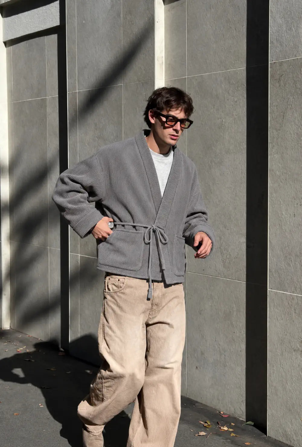 Veste kimono homme grise style cardigan portée nouée sur un pantalon beige devant un mur en pierre